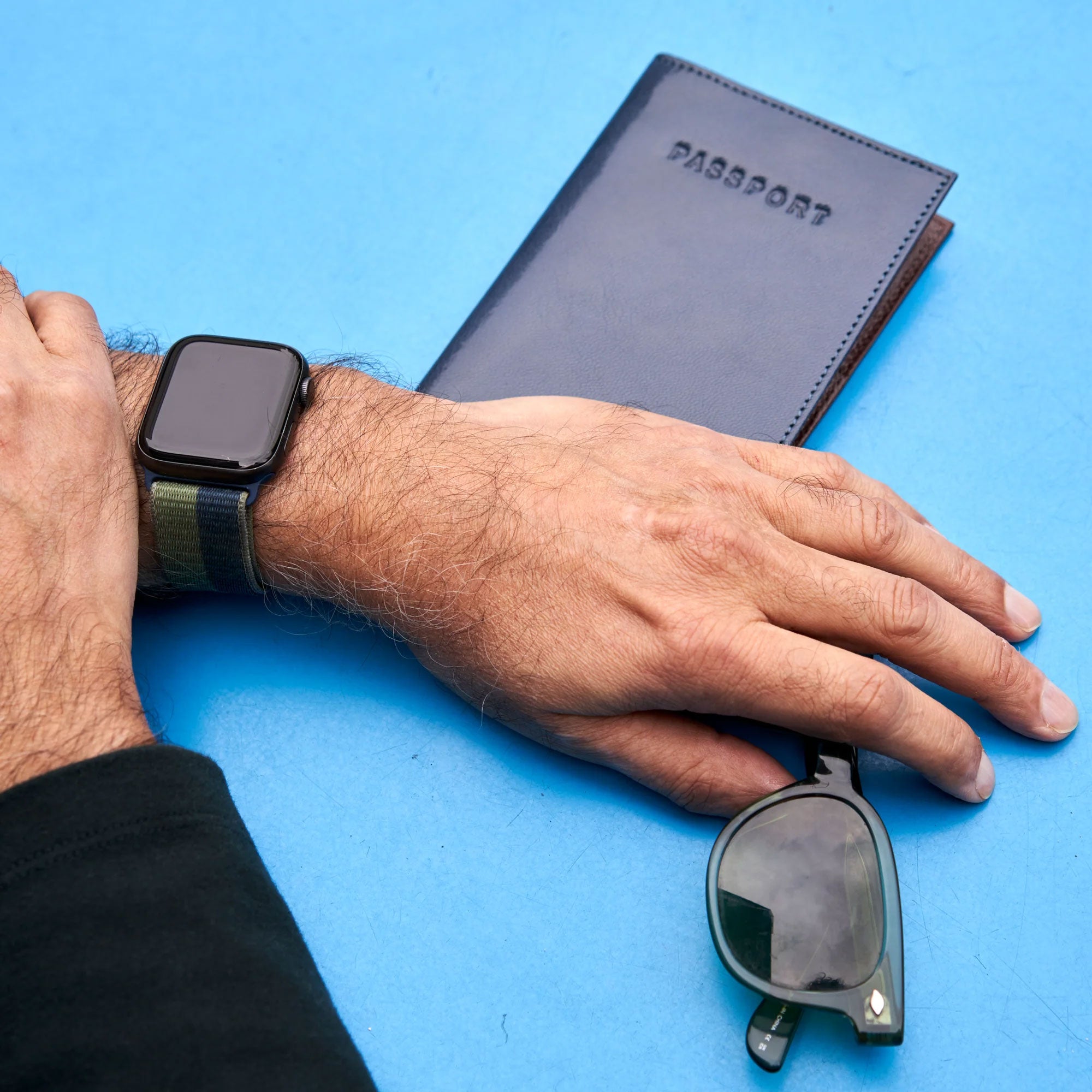 A close-up of a man's hand wearing a smartwatch, resting on a passport and a pair of sunglasses against a blue background.