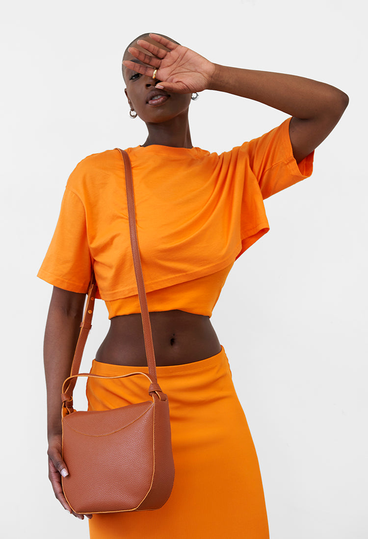 Woman model in orange outfit shielding eyes, with a brown crossbody bag, against a white backdrop