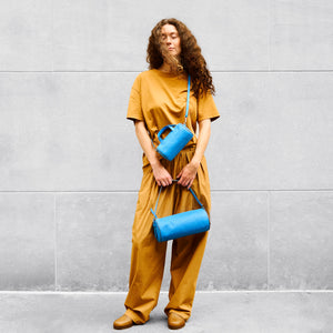 Person wearing a mustard yellow outfit holding two blue handbags against a gray wall - AZURE