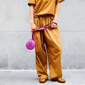 Person wearing mustard yellow outfit holding a magenta round leather bag against a gray wall -MAGENTA