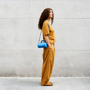 Woman in mustard yellow outfit carrying a small blue leather handbag crossbody against a gray wall - AZURE