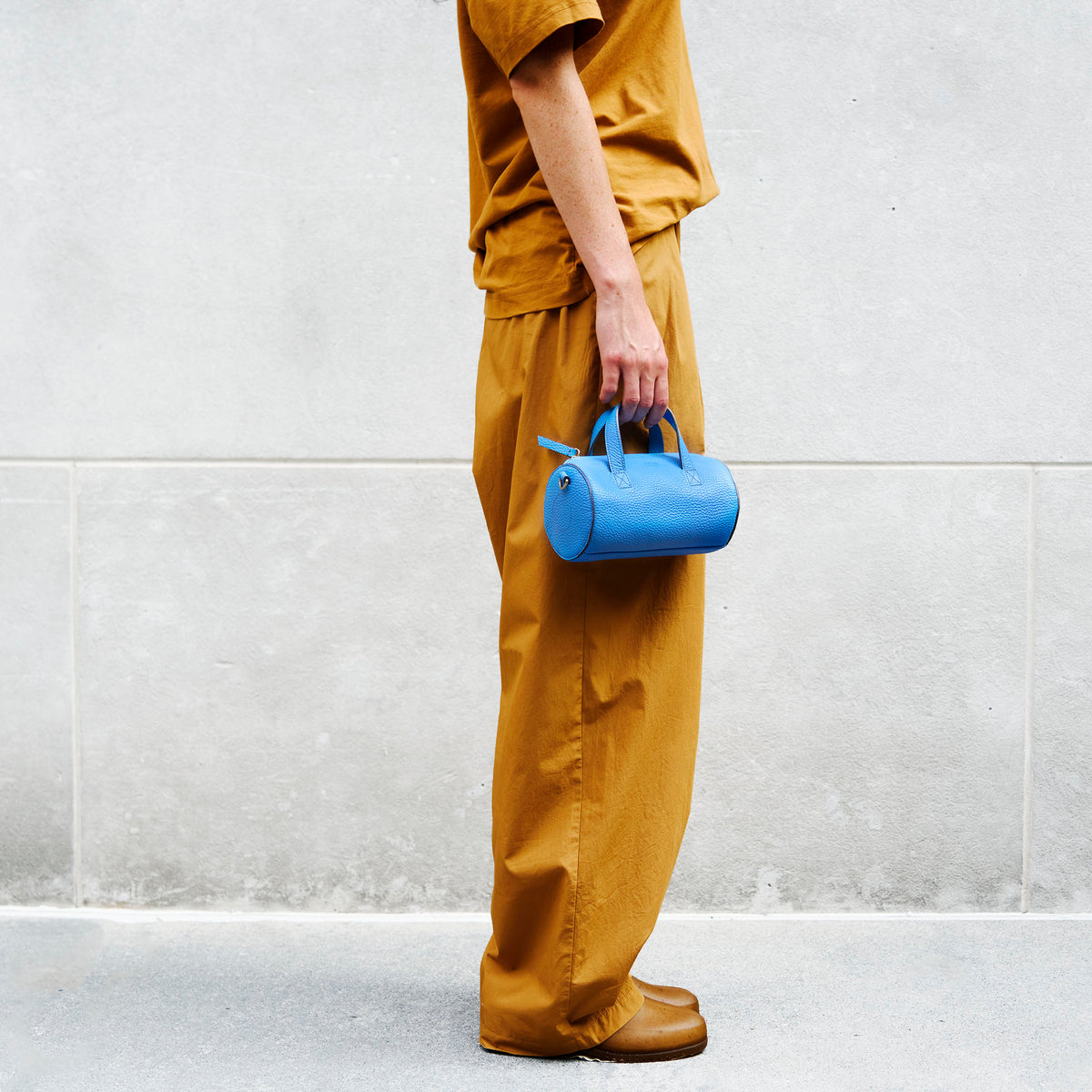 Person wearing mustard yellow outfit holding a blue leather barrel bag against a light gray wall - AZURE