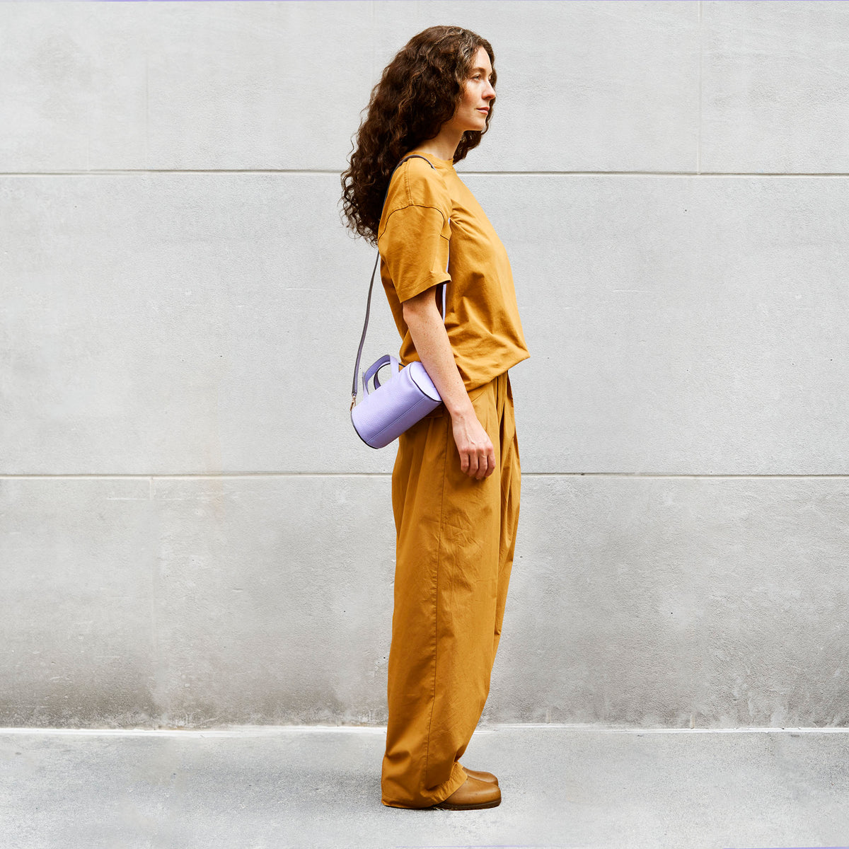 Woman in mustard yellow outfit carrying  a light purple leather bag crossbody against a gray wall - LAVENDER