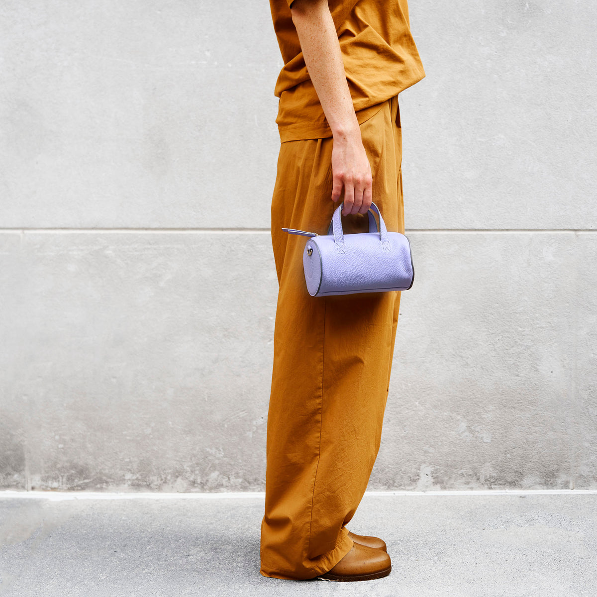 Person holding a light purple leather  handbag in hand against a gray wall - LAVENDER