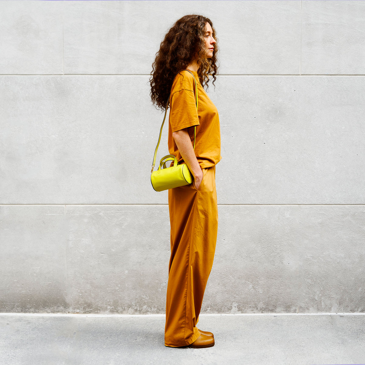 Woman in mustard yellow outfit holding a yellow leather handbag against a gray wall - LIME