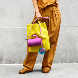 Person holding a yellow tote bag with a purple handbag attached, wearing brown pants and shoes against a gray wall - MAGENTA