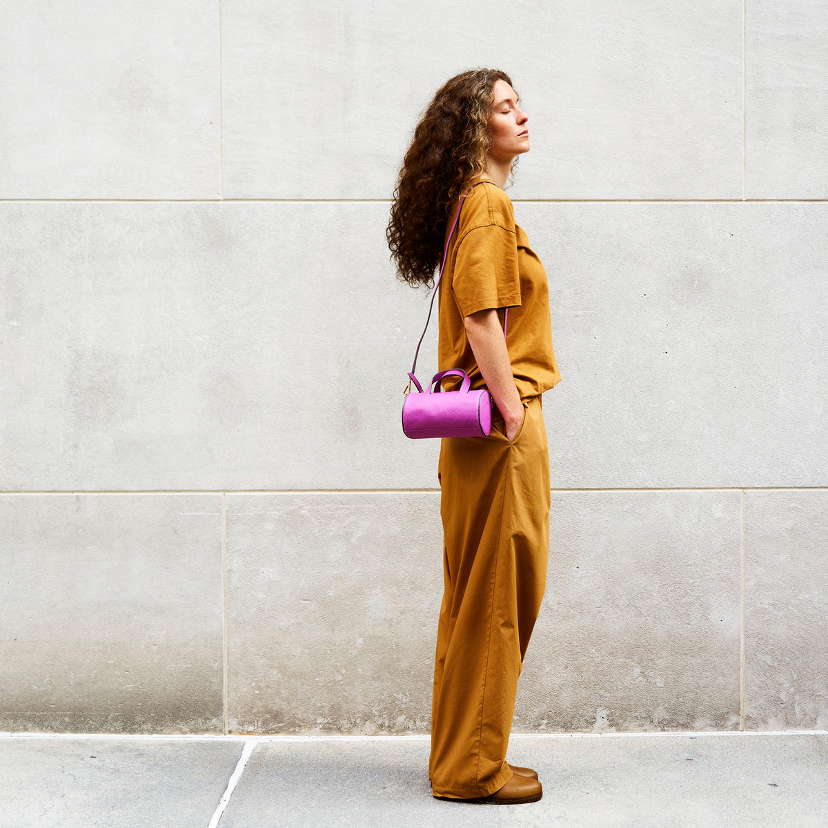 Woman in mustard yellow outfit holding a purple handbag against a light gray wall - MAGENTA