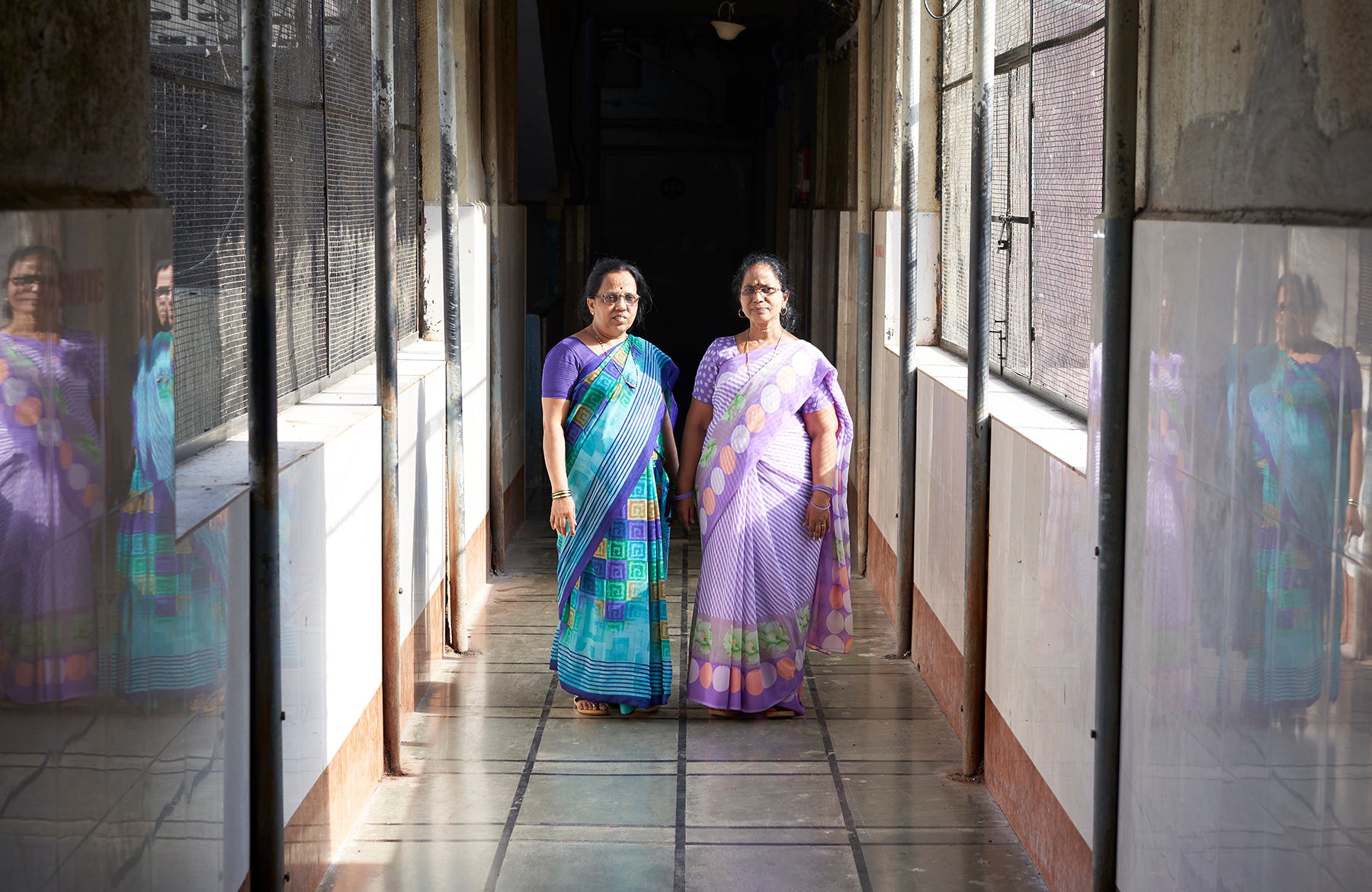 ROHINI P & ROHINI M in colorful sarees stand in a sunlit corridor with reflective walls.
