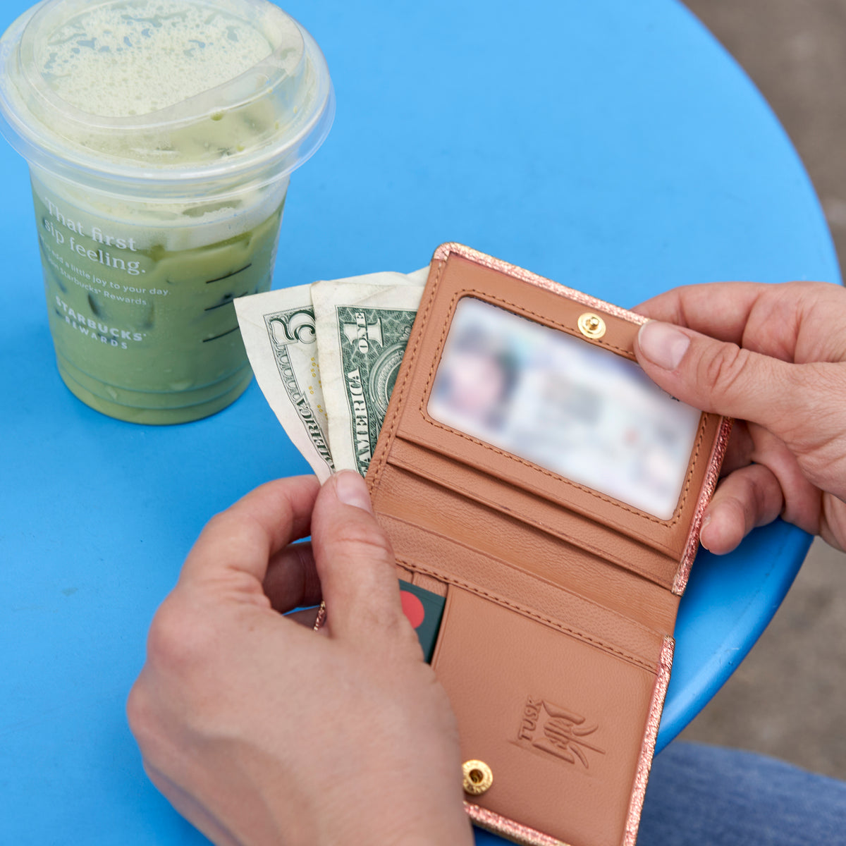 Person holding a rose gold wallet with money and a drink on a blue surface - ROSE