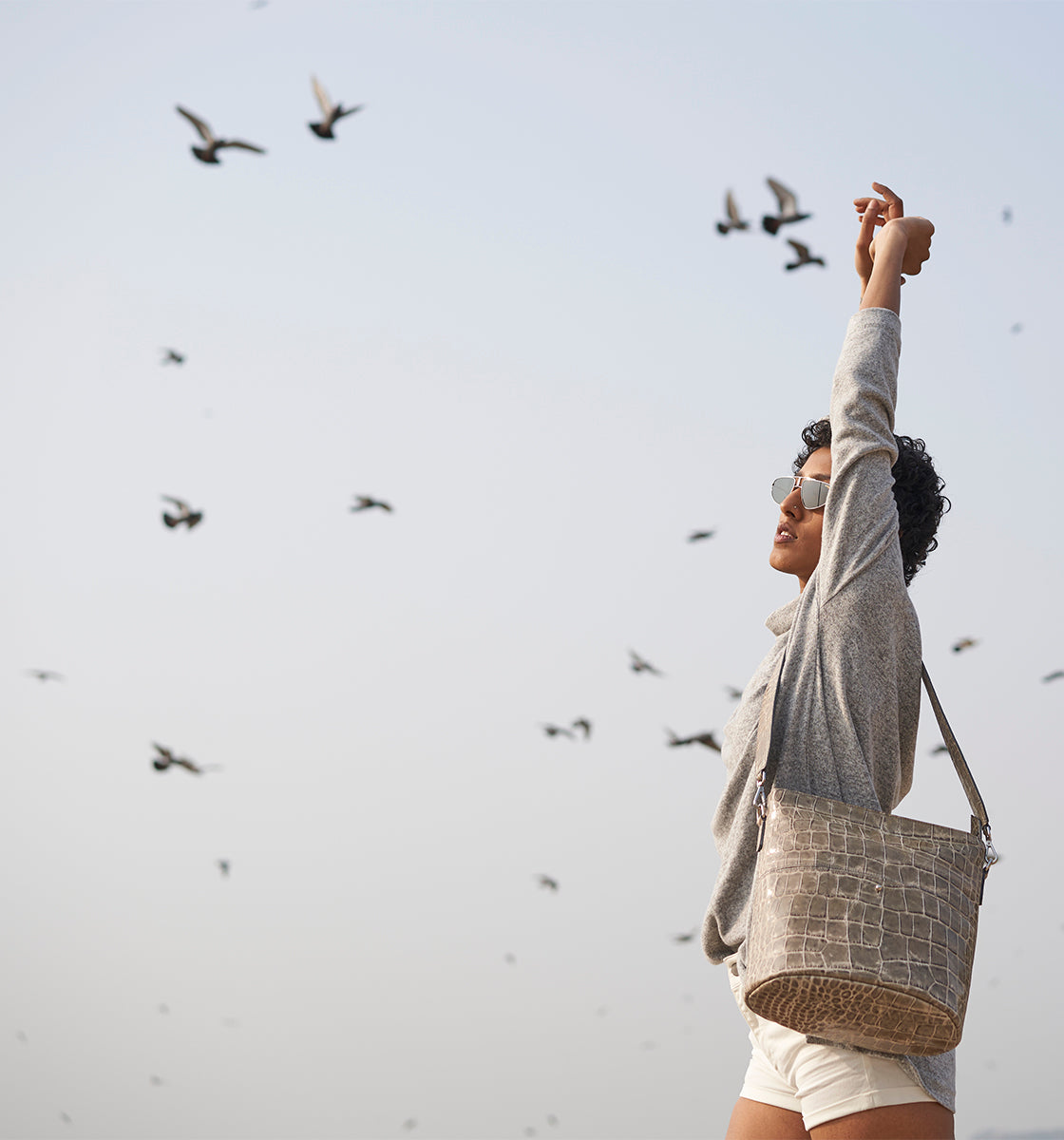 Woman stretching with arms up, wearing sunglasses and a grey top, with a crossbody bag. Birds are flying in the sky.