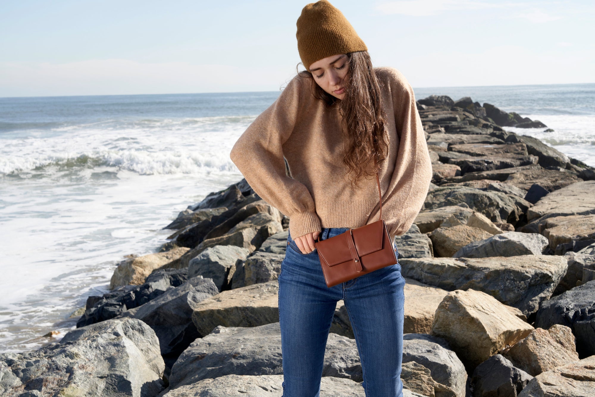 Woman in beige sweater and beanie with brown bag by the sea.