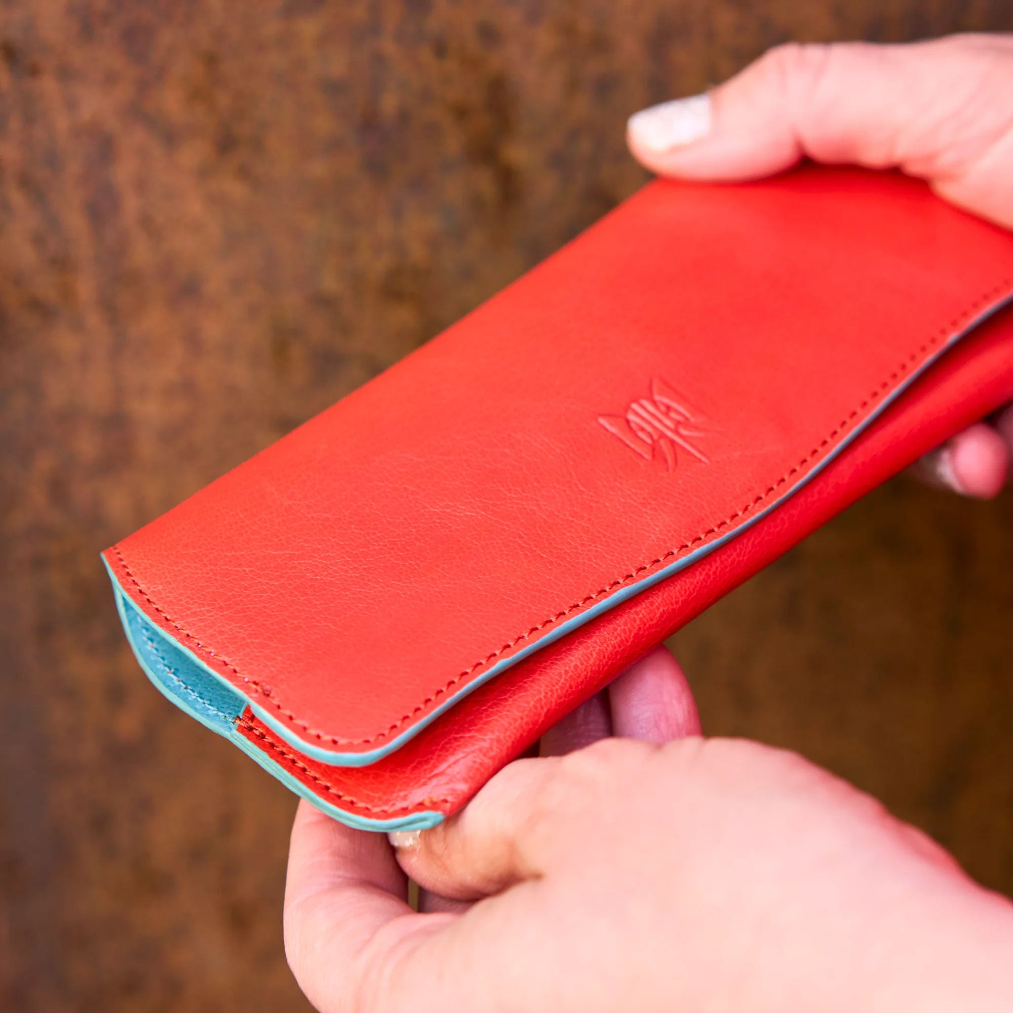 A person's hand holds a vibrant red leather wallet with teal accents, against a rustic brown background.