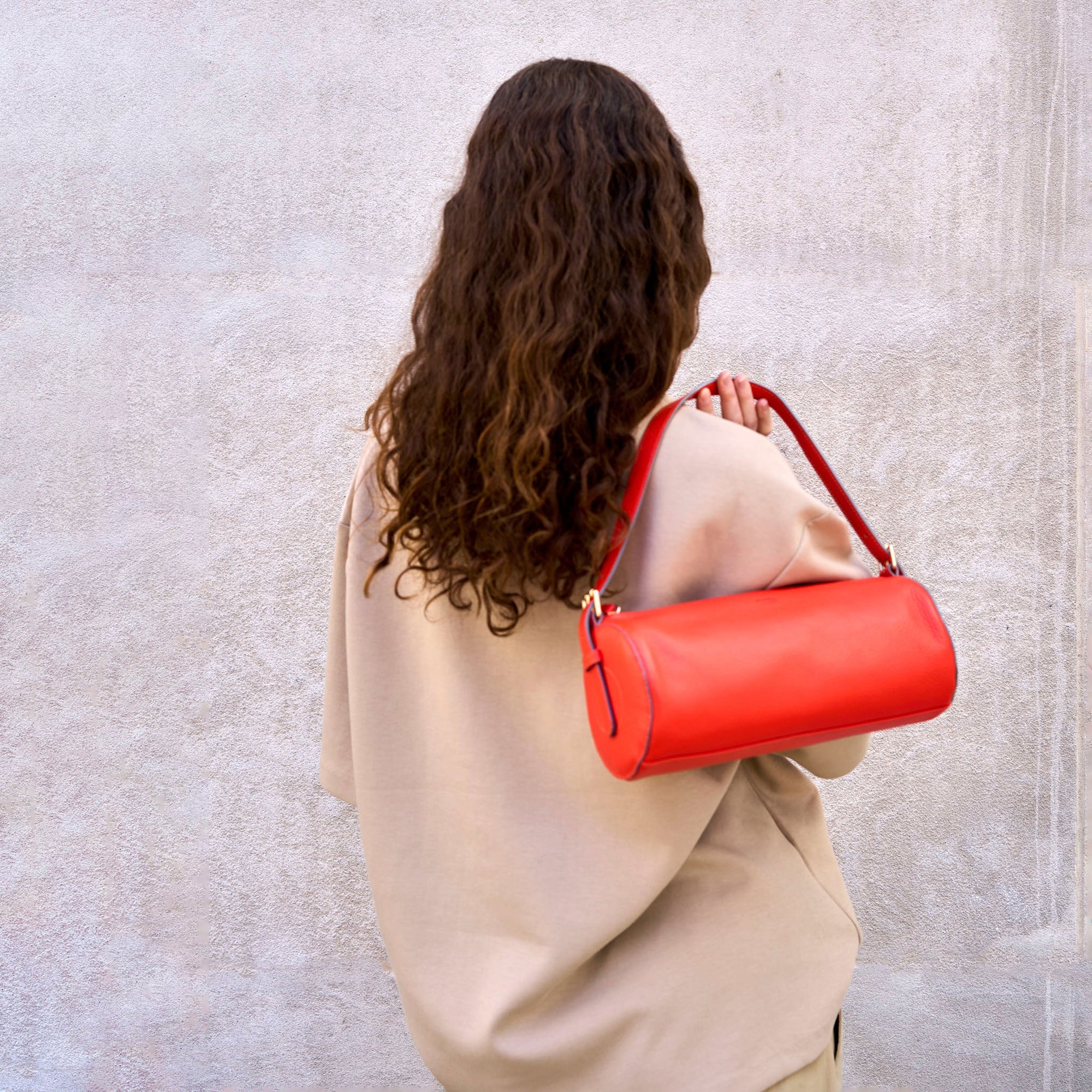 A woman with long, curly hair wears a beige oversized top and carries a vibrant red handbag over her shoulder against a textured wall.