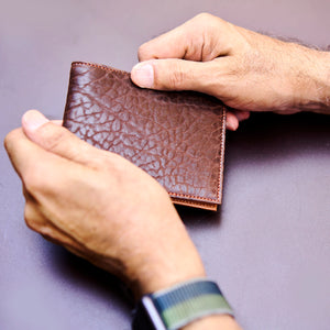 Brown leather wallet held by a person's hands on a gray background -CHOCOLATE