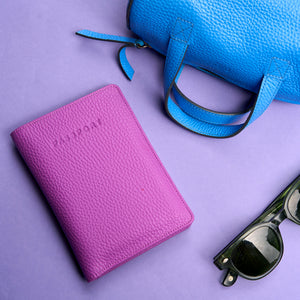 Magenta leather passport wallet, blue handbag, and sunglasses on a purple background -MAGENTA