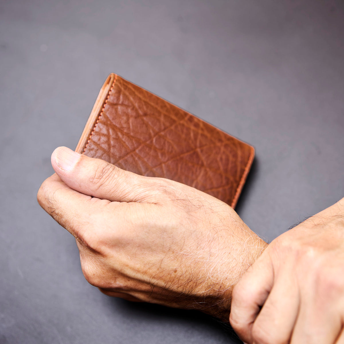 Brown leather wallet held by a hand against a gray background - TAN