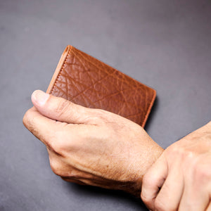 Brown leather wallet held by a hand against a gray background - TAN