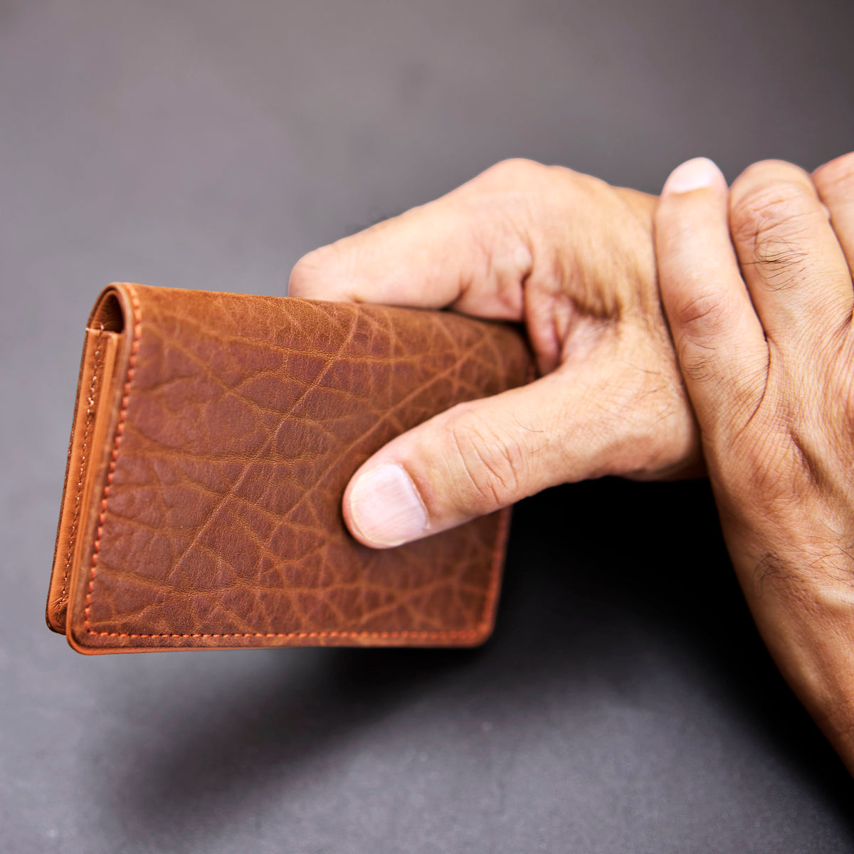 Brown leather wallet held by a hand against a gray background - TAN