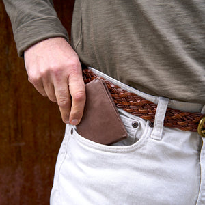 Person wearing a woven belt with a brown leather wallet in the pocket against a wooden background - BROWN