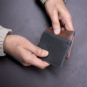 Two hands holding a leather wallet against a gray background - BROWN