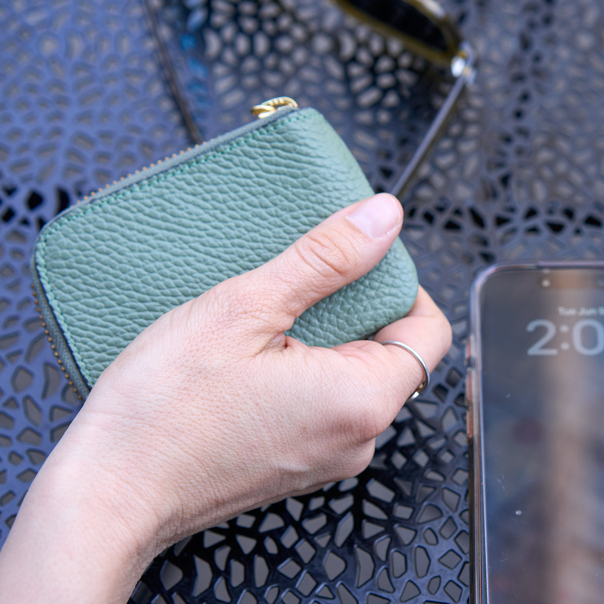 Tusk 194 Ascot small zip card wallet, held closed beside a phone on a cafe table - SAGE