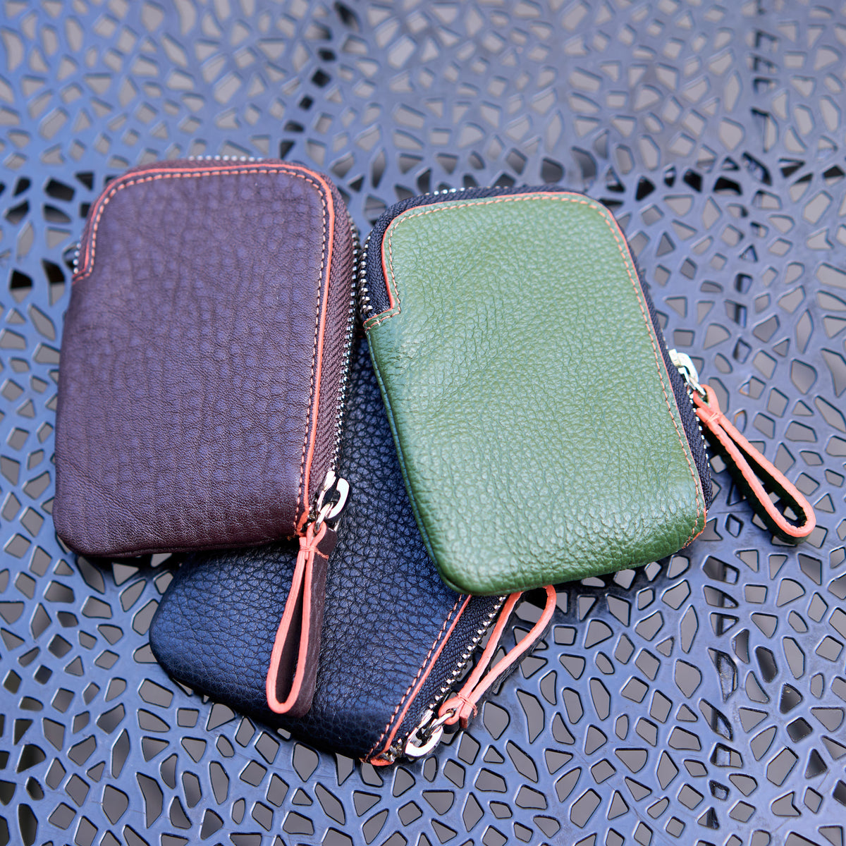 Three Amsterdam Small Zip Card Wallets in chocolate brown, forest green, and black pebbled leather, clustered together on a dark gray patterned metal tabletop. Each wallet has a curved top edge highlighted by an orange stitched border - BLACK 