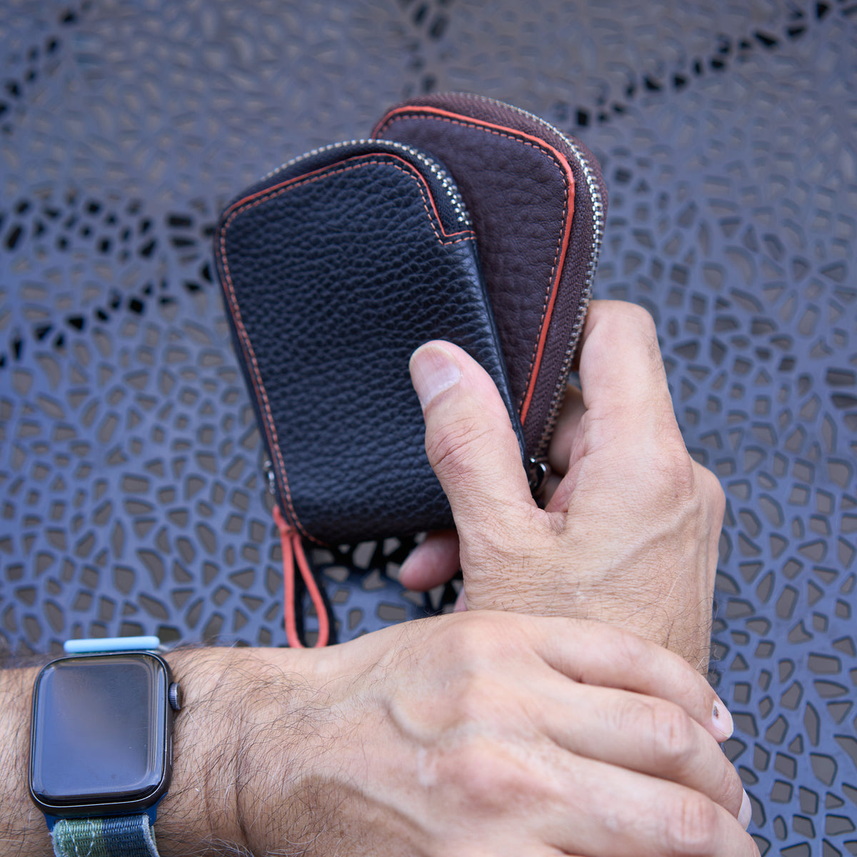 A close-up shot of a person's hands holding two Amsterdam Small Zip Card Wallets stacked together against a dark gray, textured metal patio tabletop. The front wallet is Black pebbled leather, and the one behind it is Chocolate brown pebbled leather. Both compact wallets feature a distinct curved top edge highlighted by a contrasting orange stitched border and a silver metal zipper - BLACK