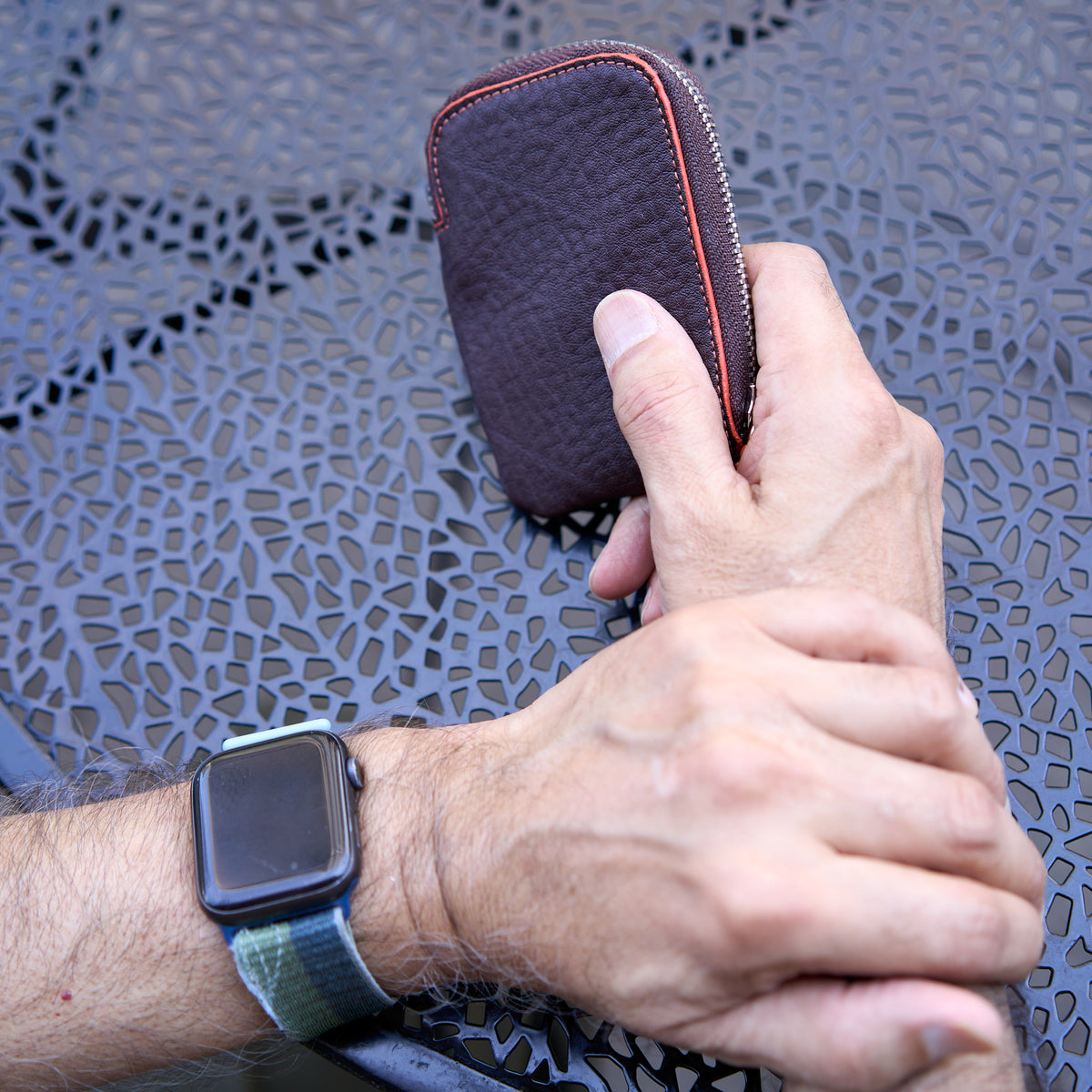 An overhead, close-up shot of a person’s hands resting on a dark gray, textured metal patio tabletop. One hand is holding an Amsterdam Small Zip Card Wallet in Chocolate brown pebbled leather, showing its curved top edge with a contrasting orange stitched border. The person's other wrist is visible, wearing an Apple Watch - CHOCOLATE