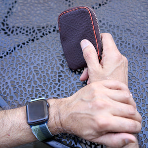 An overhead, close-up shot of a person’s hands resting on a dark gray, textured metal patio tabletop. One hand is holding an Amsterdam Small Zip Card Wallet in Chocolate brown pebbled leather, showing its curved top edge with a contrasting orange stitched border. The person's other wrist is visible, wearing an Apple Watch - CHOCOLATE
