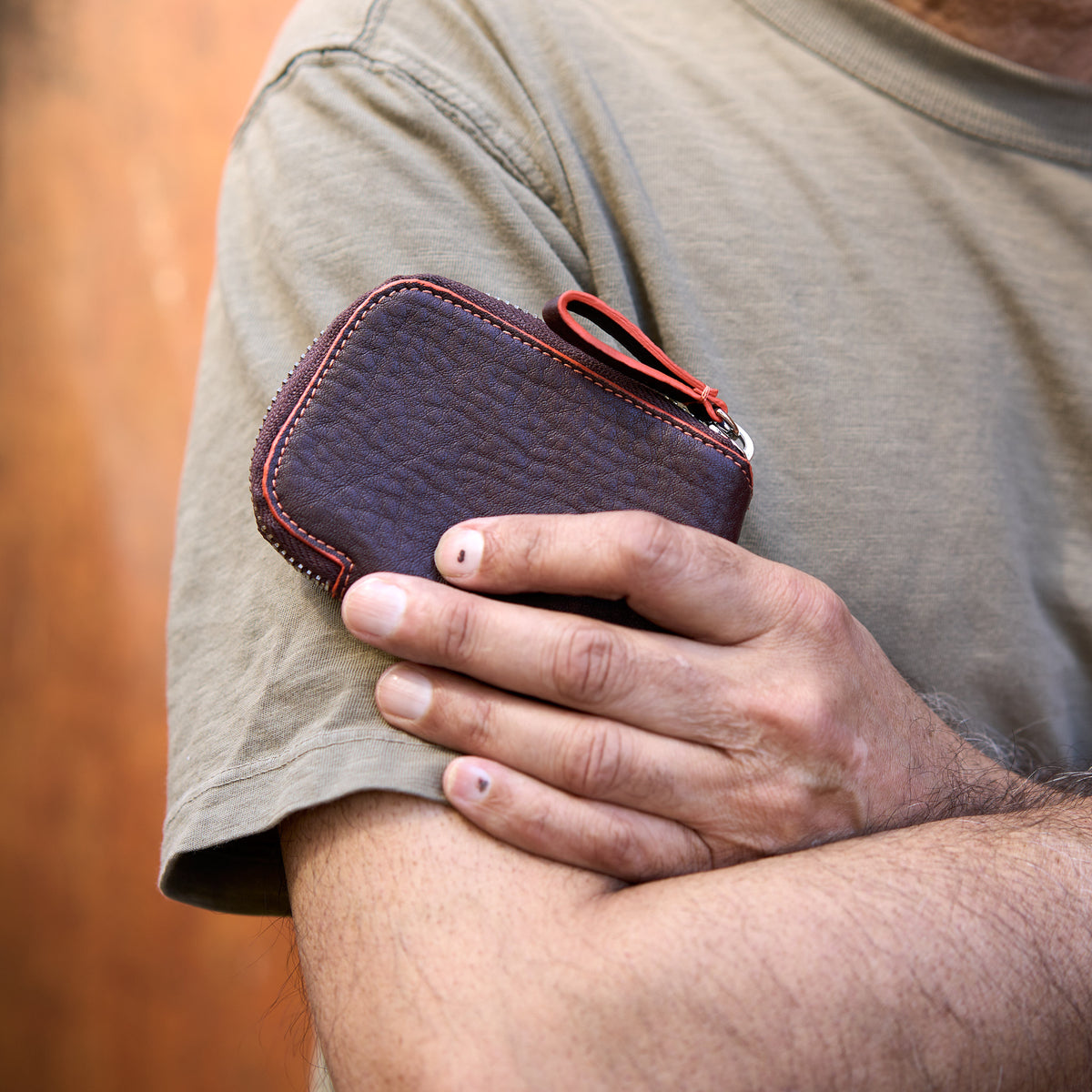 A close-up shot of a person with their arms crossed, holding a chocolate brown pebbled leather Amsterdam Small Zip Card Wallet. The wallet features a contrasting orange stitched border along its curved edge, a silver metal zipper, and an attached matching brown leather pull tab with an orange accent - CHOCOLATE