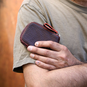 A close-up shot of a person with their arms crossed, holding a chocolate brown pebbled leather Amsterdam Small Zip Card Wallet. The wallet features a contrasting orange stitched border along its curved edge, a silver metal zipper, and an attached matching brown leather pull tab with an orange accent - CHOCOLATE