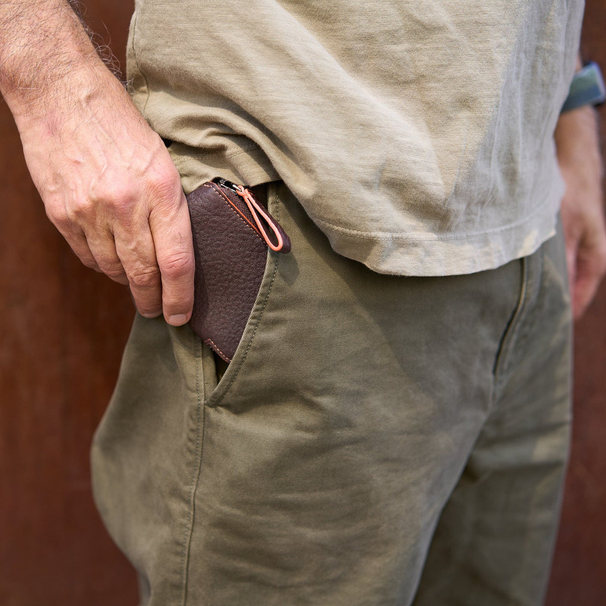 A close-up shot of a person slipping the Amsterdam Small Zip Card Wallet in Chocolate green pebbled leather into the front pocket of their olive green trousers. The compact wallet features a contrasting orange stitched edge and an orange and cream leather zipper pull loop peeking out of the pocket- CHOCOLATE
