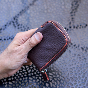 A close-up shot of a person’s hand holding the Amsterdam Small Zip Card Wallet in Chocolate brown pebbled leather. The compact wallet features a contrasting orange stitched border along its curved edge, a silver metal zipper, and an attached brown leather pull tab with an orange accent. The background shows a dark gray, textured metal patio tabletop - CHOCOLATE