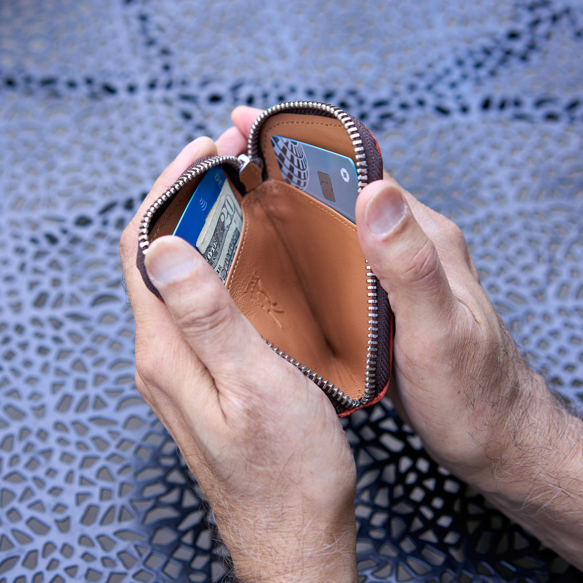 An overhead, close-up shot of a person’s hands holding open the Amsterdam Small Zip Card Wallet in Chocolate brown pebbled leather. The wallet is unzipped, revealing its smooth tan leather interior. Inside, a blue credit card is visible in a left slot behind a folded $20 bill, and a gray credit card is tucked into a slot on the right side. The silver zipper track outlines the curved top edge of the wallet, and the background shows a dark gray, patterned cutout metal tabletop - BLACK