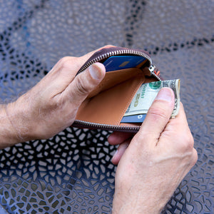A close-up shot of a person’s hands holding the Amsterdam Small Zip Card Wallet in Chocolate brown pebbled leather. The wallet features a contrasting orange stitched border along its curved edge, a silver metal zipper, and an attached brown leather pull tab. The background shows a dark gray, textured metal patio tabletop - CHOCOLATE