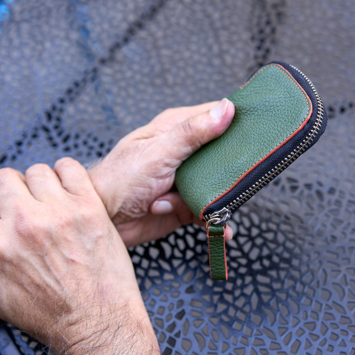 close-up shot of a person’s hands holding the Amsterdam Small Zip Card Wallet in Forest green pebbled leather. The wallet features a contrasting orange stitched border along its curved edge, a silver metal zipper, and an attached matching green leather pull tab. The background shows a dark gray, textured metal patio tabletop - FOREST