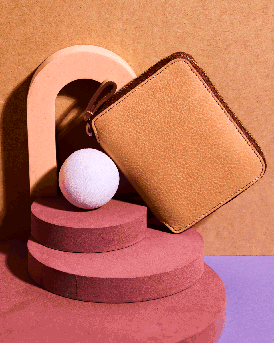 A tan leather wallet displayed on a geometric arrangement of pink and beige shapes with a white spherical object.