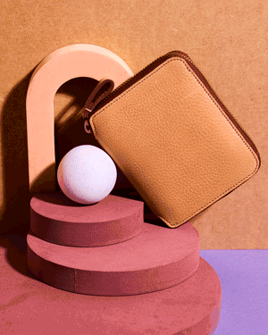 6 of 6: A tan leather wallet displayed on a geometric arrangement of pink and beige shapes with a white spherical object.