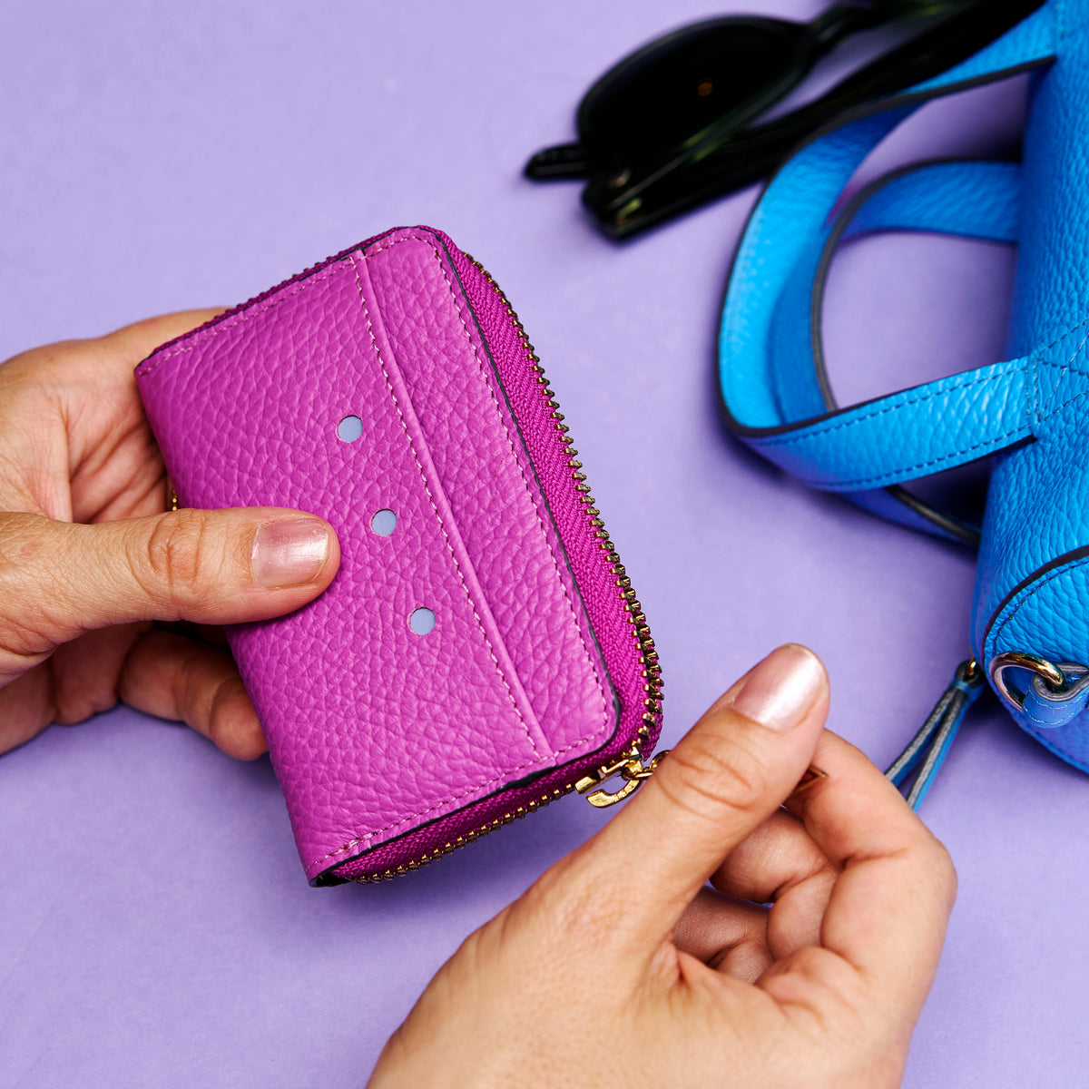 Purple wallet held by a hand with a blue bag in the background on a purple surface - MAGENTA