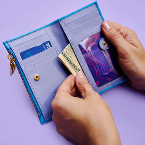 Person holding an open blue leather wallet with a  light blue interior on a light purple background -AZURE