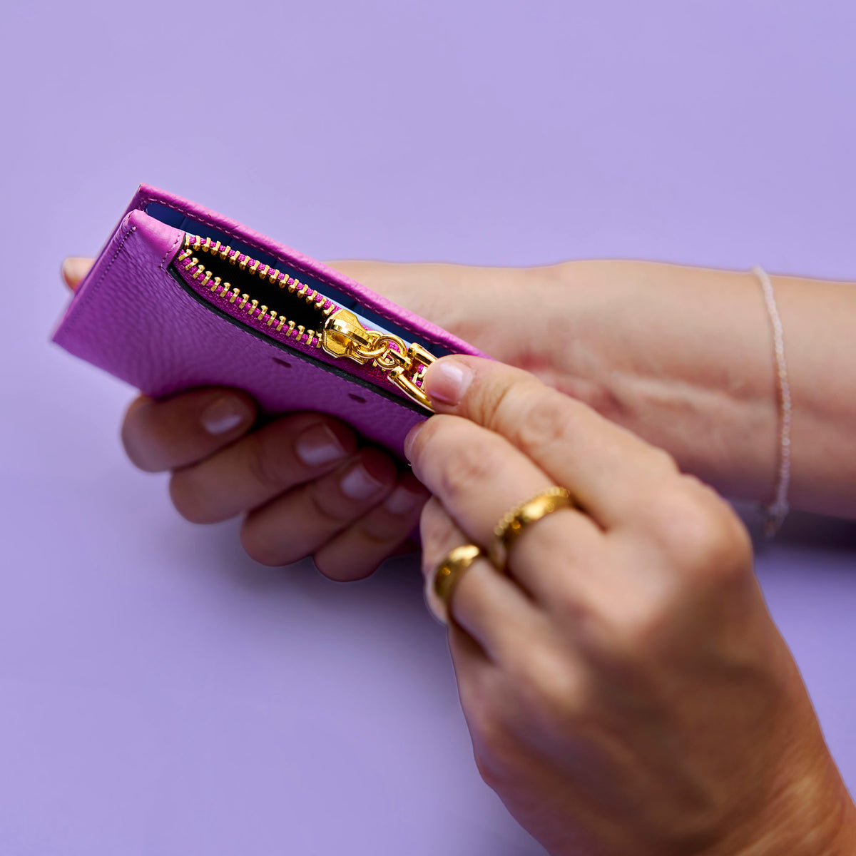 Hand holding a small magenta leather wallet with gold zipper against a light purple background - MAGENTA