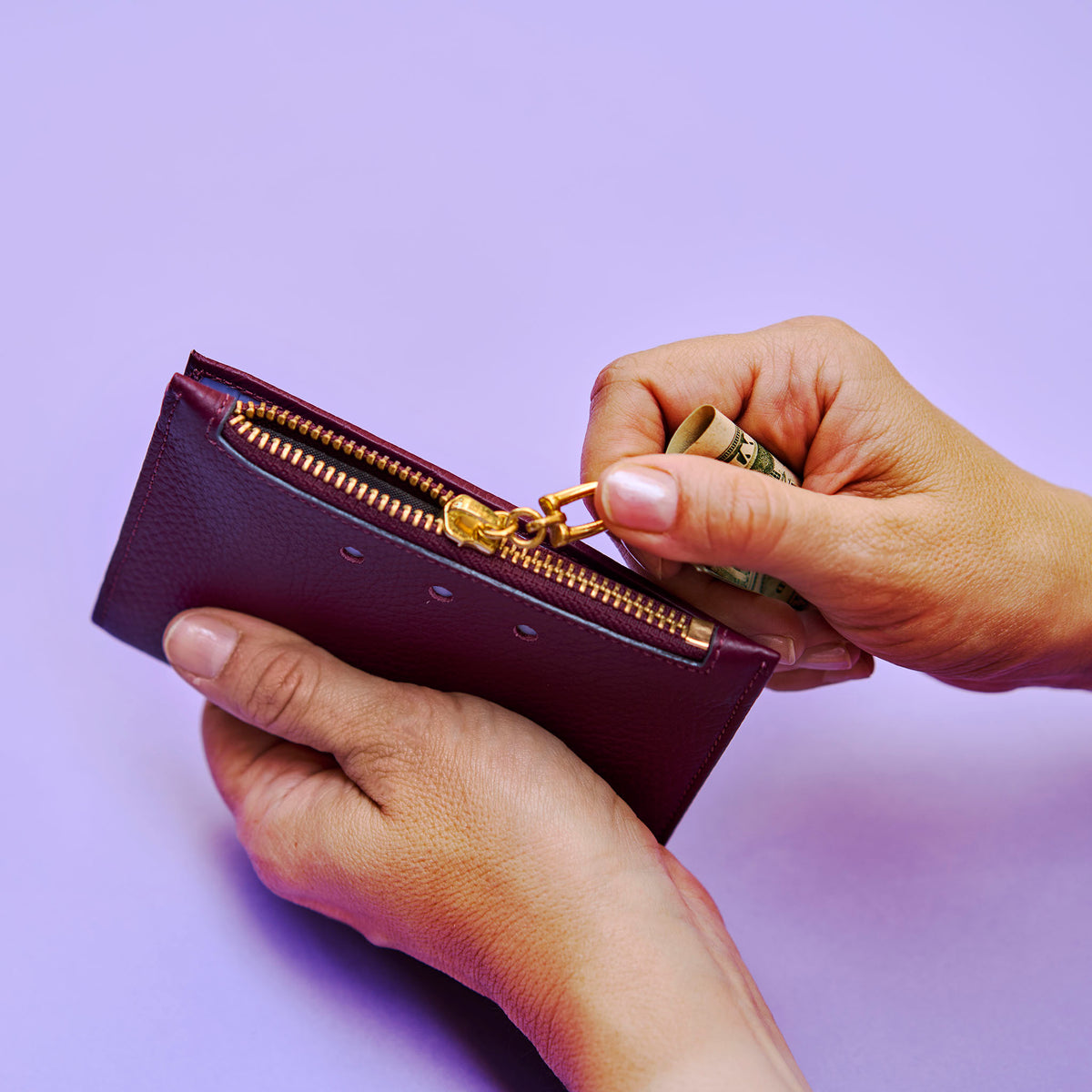 Person opening a purple wallet with a gold zipper on a light purple background - OXBLOOD