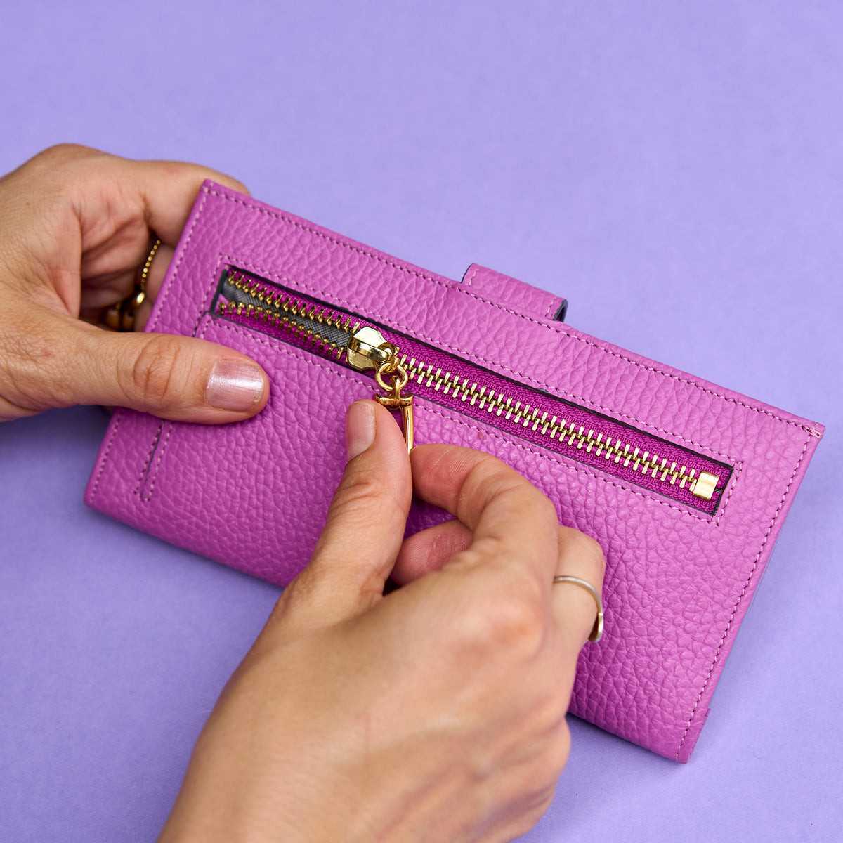 Magenta wallet with gold zipper being opened by hands on a purple background - MAGENTA