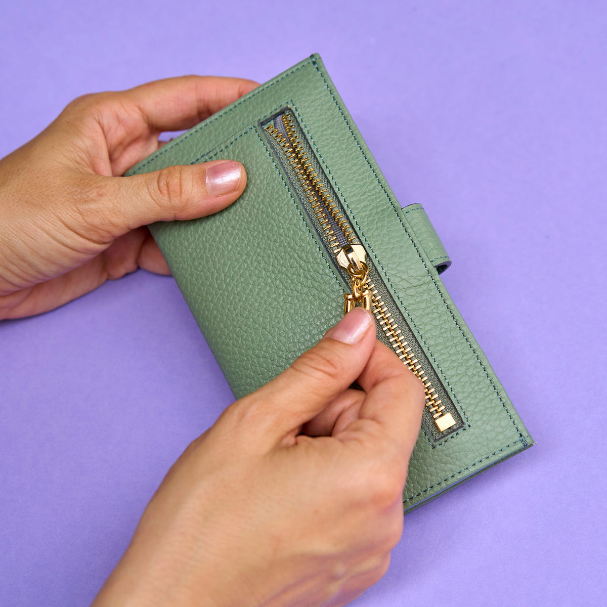 Green wallet with gold zipper held by hands against a purple background - SAGE