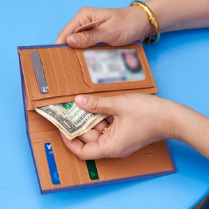 A person opening a metallic blue leather slim wallet with a tan interior, showing card slots and cash, against a bright blue background - NAVY