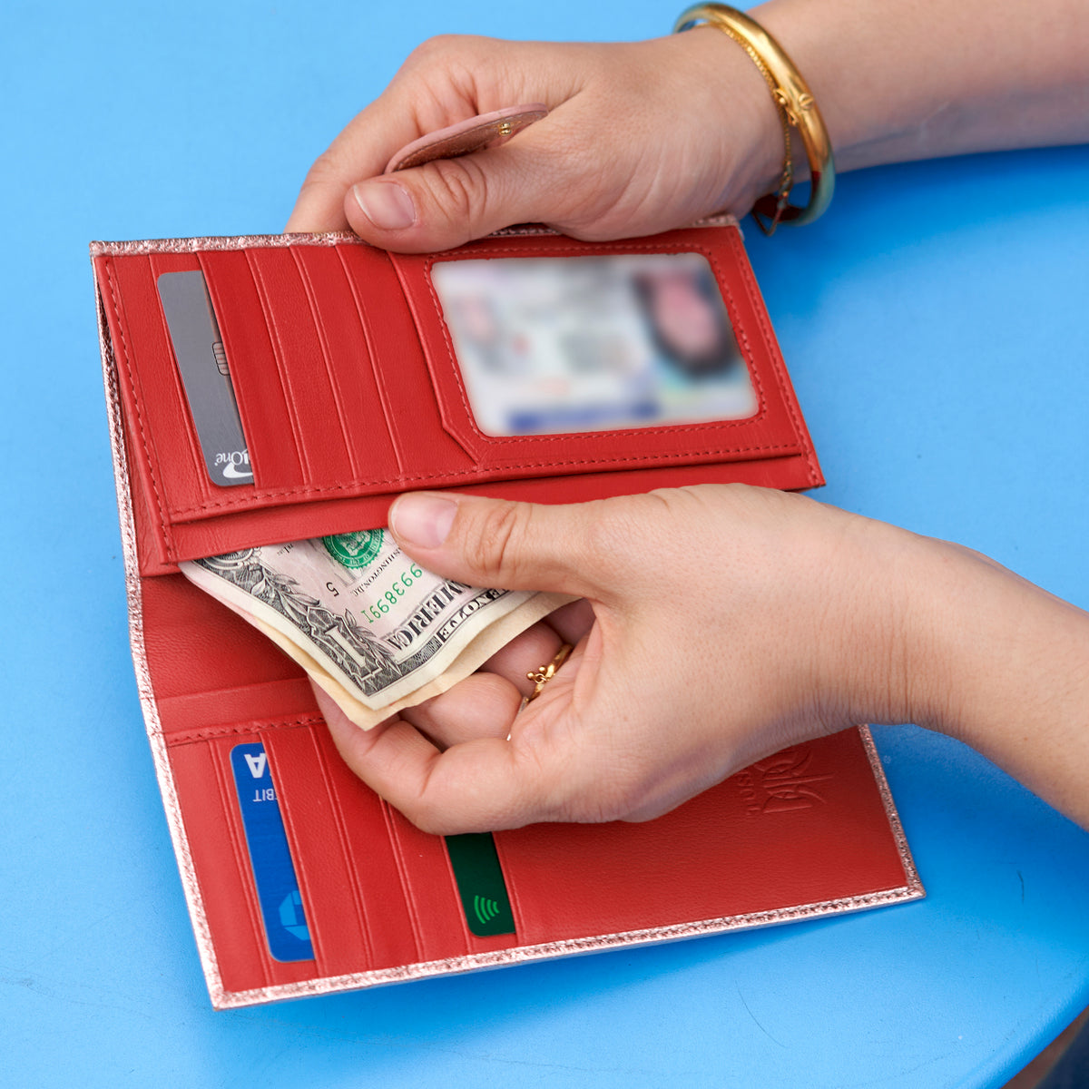 Open metallic rose gold wallet with bright red leather interior, card slots, cash compartment, and ID window held over a bright blue table with gold jewelry accents. - ROSE GOLD