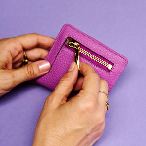 Magenta leather wallet with gold zipper held by hands against a purple background - MAGENTA