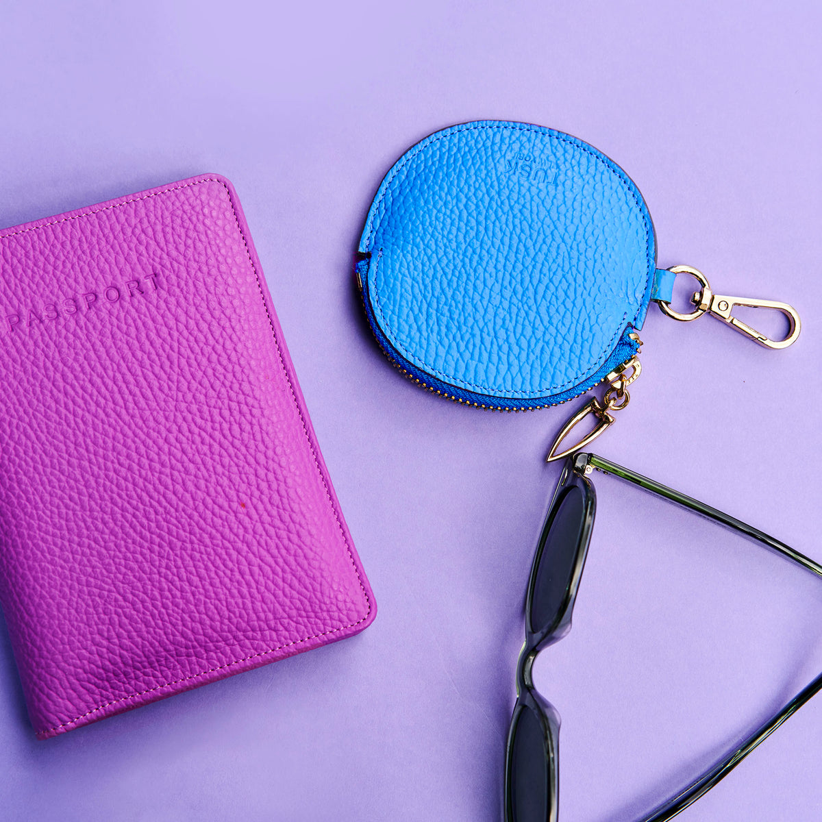 Magenta leather passport holder, blue leather coin purse with dog-clip, and sunglasses on a purple background - AZURE