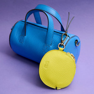 Blue leather barrel handbag with a yellow round pouch charm attached on a purple background - LIME