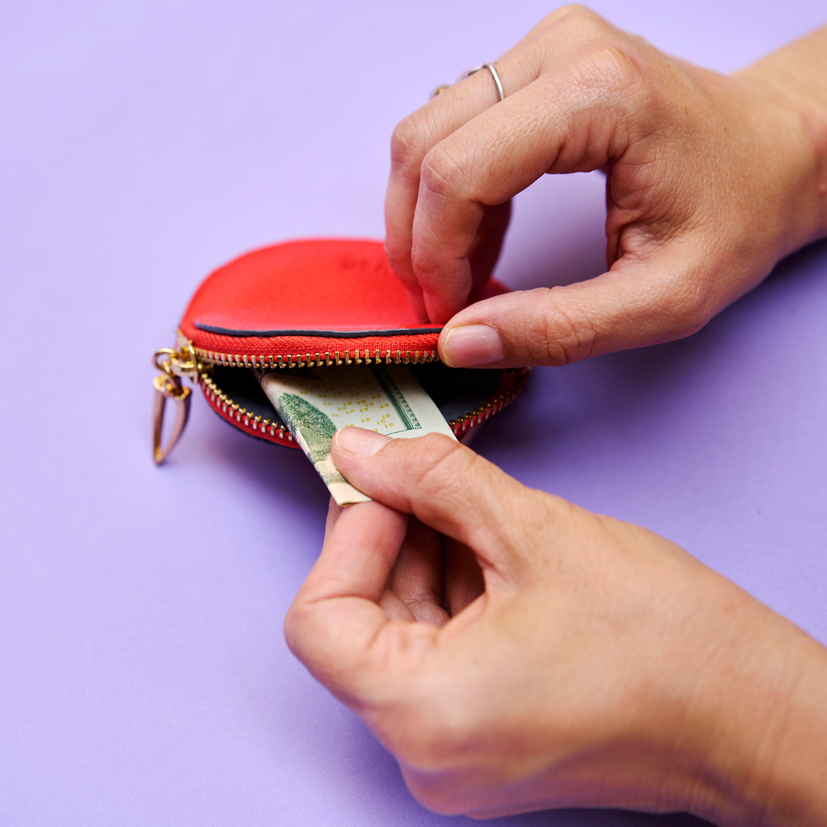 Person opening an orange round bag charm to take out a banknote on a purple background - ORANGE